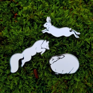 Op de afbeelding: Drie witte emaille pinnen met daarop een vos, een konijn en een opgerold konijn. De pinnen zijn op een groene mossige achtergrond.
