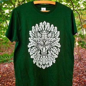 Op de afbeelding: Een donkergroen T-shirt met een witte grafische print van een wolvenkop omringd door eikenbladeren. De tekst "Art of Mäquenda" is gedrukt op de onderkant van het shirt.