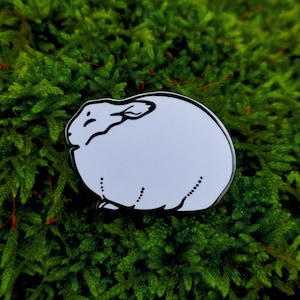 Op de afbeelding: Een witte emaille pin in de vorm van een konijn, met zwarte omtrek en details. De pin is geplaatst tegen een achtergrond van levendig groen mos. Een schattig accessoire.