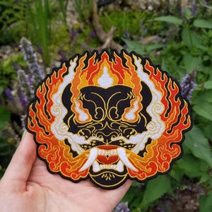 Op de afbeelding: Geborduurde patch met een gestileerd demonengezicht met vlammen. Het ontwerp omvat een zwart gezicht met gouden details, witte hoektanden en een rode mond. De vlammen zijn in oranje, geel en rood, met een zwarte rand. De patch wordt in een hand gehouden tegen een achtergrond van groen gebladerte.