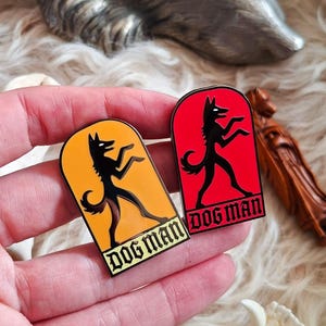 Puede incluir: Dos pines de esmalte con una figura negra con forma de perro en posición de caminar. Uno tiene fondo naranja, el otro rojo. Ambos pines tienen las palabras "DOG MAN" en fuente gótica. Los pines se sostienen en una mano.
