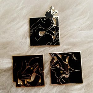 Op de afbeelding: Drie zwarte en gouden emaille pinnen met gestileerde zwarte wolfsilhouetten. De pinnen zijn vierkant en hebben een gouden rand.
