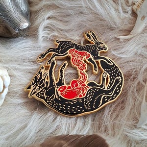 Op de afbeelding: Een zwarte en gouden emaille pin met een gestileerd ontwerp van twee wolven en een konijn. Het konijn springt boven de wolven, met een rood bloemenontwerp ertussen. De pin heeft een gouden rand en witte stipdetails.