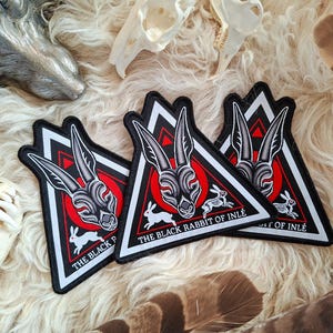 Op de afbeelding: Drie zwart, rood en wit geborduurde patches met een konijnontwerp. Elke patch heeft een konijnenkop in een rode cirkel, een klein rennend konijn en de tekst "THE BLACK RABBIT OF INLÉ".