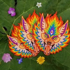 Op de afbeelding: Geborduurde patches met een gestileerde wolf met een vurige, regenboogkleurige manen en een derde oog. De patches zijn geplaatst op een groot groen blad, met kleurrijke bloemen verspreid.