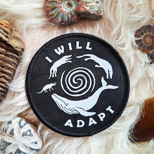 以下が含まれることがあります： 白のテキストとイラストが入った黒い円形のパッチ。テキストは、中央のらせん状のデザインの周りに「I WILL ADAPT」と書かれています。イラストには、クジラ、小さな動物、その他の2つの生き物が含まれています。パッチはさまざまな化石に囲まれています。