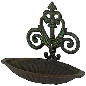 May include: Ornate, cast iron wall-mounted soap dish with a fleur-de-lis design. The dish has a dark, weathered finish with green highlights. The decorative bracket features scrolled accents and a central vertical element.