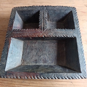 May include: A dark wooden divided tray with four compartments. The tray has a carved, textured surface and a decorative border. The wood has a natural, aged appearance, with variations in colour and grain. The tray is likely used for organising small items.