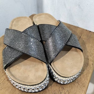 Puede incluir: Un par de sandalias negras con un dise&ntilde;o de tiras cruzadas. Las tiras est&aacute;n cubiertas de pedrer&iacute;a y las sandalias tienen una suela de plataforma con un ribete de cuerda.