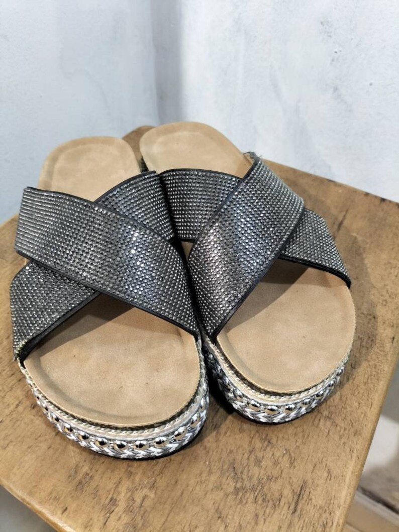 Puede incluir: Un par de sandalias negras con un dise&ntilde;o de tiras cruzadas. Las tiras est&aacute;n cubiertas de pedrer&iacute;a y las sandalias tienen una suela de plataforma con un ribete de cuerda.