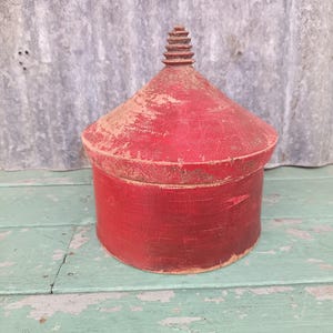 May include: A cylindrical wooden container with a conical lid featuring a tiered finial at the top. The object is finished in a distressed red paint, showing visible wear and texture across the surface of both the lid and the base.