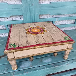 May include: A hand-painted wooden stool with a floral design. The stool has a light brown base with a red and gold trim. The top of the stool is painted with a white background and a yellow, green and red flower design.