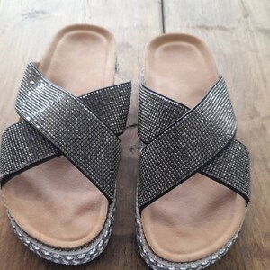 Puede incluir: Un par de sandalias negras con un dise&ntilde;o de correa cruzada. Las correas est&aacute;n cubiertas de diamantes de imitaci&oacute;n.