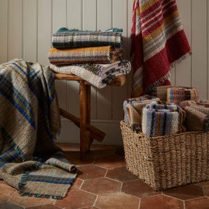 May include: A selection of folded and draped woven blankets and throws in various colours and patterns. Some are stacked on a wooden stool, others are in a wicker basket, and one is draped over a wall. Colours include red, blue, green, and yellow.