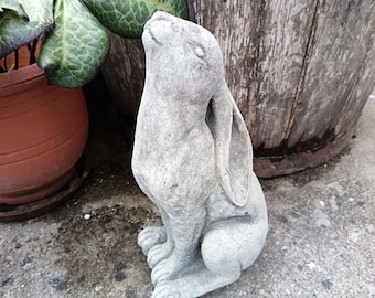 Könnte beinhalten: Eine graue Betonkaninchenstatue mit nach oben geneigtem Kopf. Das Kaninchen hat lange Ohren und sitzt auf seinen Hinterbeinen. Die Statue steht auf einer Betonoberfläche, im Hintergrund ein Blumentopf und ein Holzfass.