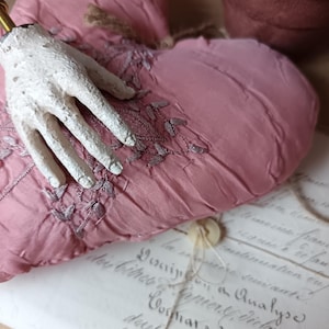 May include: A dusky pink heart-shaped cushion with embroidered detailing and a white hand sculpture. The cushion rests on vintage paper with handwritten script and a button.