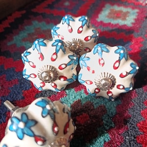 May include: Four white ceramic cabinet knobs with a floral design in blue and red. Each knob has a silver-coloured metal base and screw. The knobs are arranged on a colourful patterned rug.