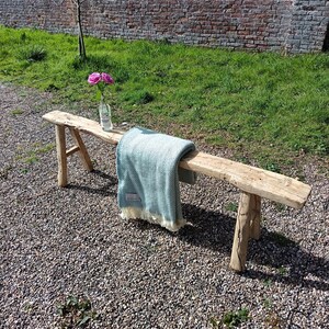 May include: A weathered wooden bench with a light blue and white blanket draped over it. The bench is made of untreated wood and has a natural, aged look. A glass bottle with pink flowers sits on the bench. The bench is outdoors on a gravel surface.