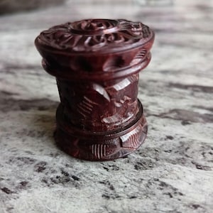 May include: A dark brown wooden box with intricate carvings. The box is cylindrical in shape and has a lid with a raised design.