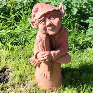May include: A terracotta garden gnome sculpture with a pointed hat, sitting with its legs crossed and feet tucked in. The gnome has a friendly face and is painted in a reddish-brown colour.