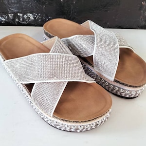May include: A pair of white platform sandals with a criss-cross strap design. The straps are covered in sparkling rhinestones.