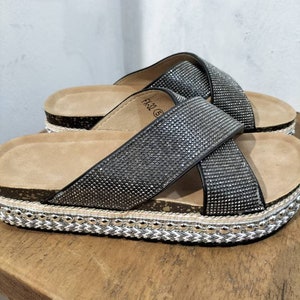 Puede incluir: Sandalias negras con una tira cruzada adornada con pedrer&iacute;a. Las sandalias tienen una suela de plataforma de corcho con un ribete trenzado.