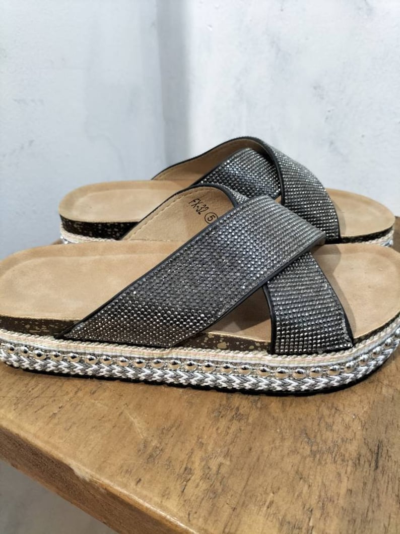 Puede incluir: Sandalias negras con una tira cruzada adornada con pedrer&iacute;a. Las sandalias tienen una suela de plataforma de corcho con un ribete trenzado.