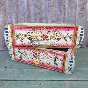 May include: Two rectangular wooden drawers with a distressed white and pink finish. Each drawer front features a hand-painted floral arrangement in a vase, accented with gold borders and blue dots along the side edges of the wooden frame.