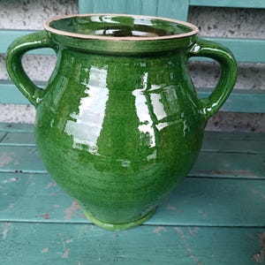 May include: A large, green ceramic pot with two handles. The pot has a glossy finish and a wide opening with a light-coloured rim. The pot is sitting on a weathered, turquoise-coloured wooden surface.