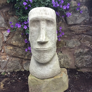 May include: A white stone sculpture of a face with a neutral expression. The sculpture is on a stone base and surrounded by purple flowers.