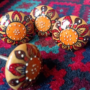 May include: Four decorative cabinet knobs with a floral design. The knobs have a tan base, orange centre, and red and brown floral accents. White dots are scattered throughout the design. The knobs are on a colourful patterned rug.