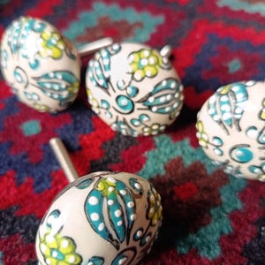 May include: Four decorative ceramic cufflinks with a cream base. Each cufflink features a hand-painted design with teal, yellow, and white floral and leaf patterns. The cufflinks have silver metal posts.