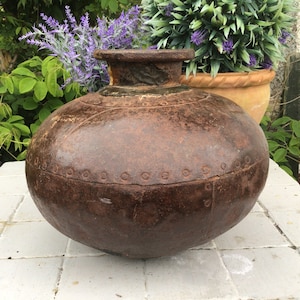 May include: A large, round, antique brown metal pot with a wide base and a narrow neck. The pot has a textured surface with decorative rivets around the middle. The pot is outdoors, with greenery in the background.