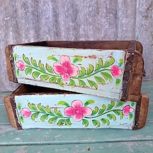 May include: Two rectangular wooden boxes stacked on top of each other. Each box features a light blue painted exterior with a hand-painted floral design consisting of bright pink flowers and green leaves. The edges are reinforced with dark brown wood.
