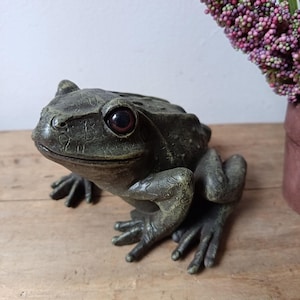 May include: A dark green frog figurine with a textured, weathered surface and prominent, dark reddish-brown eyes. The frog is depicted in a seated, alert posture with its front legs extended forward and its back legs tucked underneath its body.