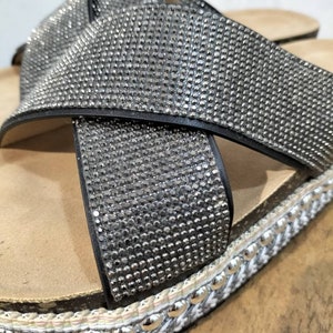 Puede incluir: Primer plano de una sandalia negra con dise&ntilde;o de tiras cruzadas. La sandalia est&aacute; cubierta de pedrer&iacute;a y tiene un ribete trenzado blanco y gris con tachuelas plateadas.