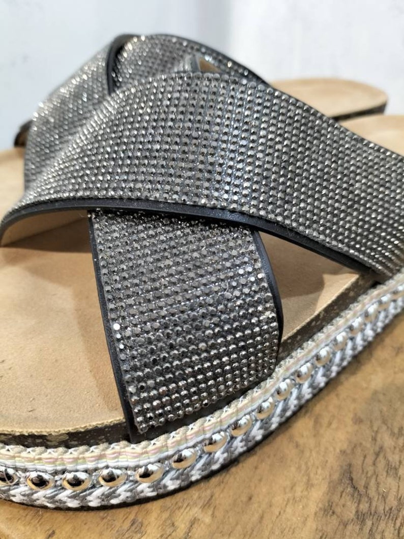Puede incluir: Primer plano de una sandalia negra con dise&ntilde;o de tiras cruzadas. La sandalia est&aacute; cubierta de pedrer&iacute;a y tiene un ribete trenzado blanco y gris con tachuelas plateadas.