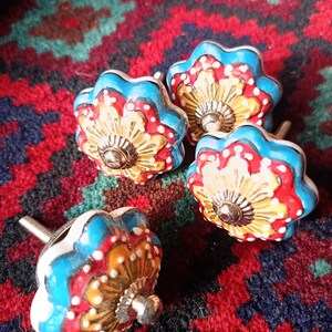 May include: Four decorative ceramic cabinet knobs. Each knob has a floral design with blue, red, yellow, and white detailing. The knobs have a silver-coloured metal base and screw.