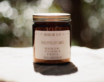 May include: A brown glass jar candle with a gold lid. The label reads "Arrows and Olive Eucalyptus Soy Candle" and includes details about the candle, such as the burn time and net weight. The candle is sitting on a light brown fabric surface.