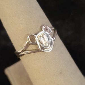 Puede incluir: Un anillo de tono plateado que presenta un motivo central con forma de cabeza de rat&oacute;n de dibujos animados con dos orejas redondeadas y una cara sonriente. El aro del anillo es fino y met&aacute;lico, con un acabado pulido que refleja la luz.