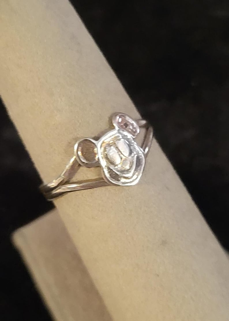 Puede incluir: Un anillo de tono plateado que presenta un motivo central con forma de cabeza de rat&oacute;n de dibujos animados con dos orejas redondeadas y una cara sonriente. El aro del anillo es fino y met&aacute;lico, con un acabado pulido que refleja la luz.