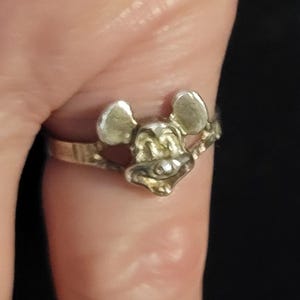 Puede incluir: Un anillo plateado con un dise&ntilde;o de la cara de Mickey Mouse. El anillo tiene una banda delgada, y la cara de Mickey Mouse est&aacute; detallada con orejas, nariz y una boca sonriente. El anillo se lleva en un dedo.