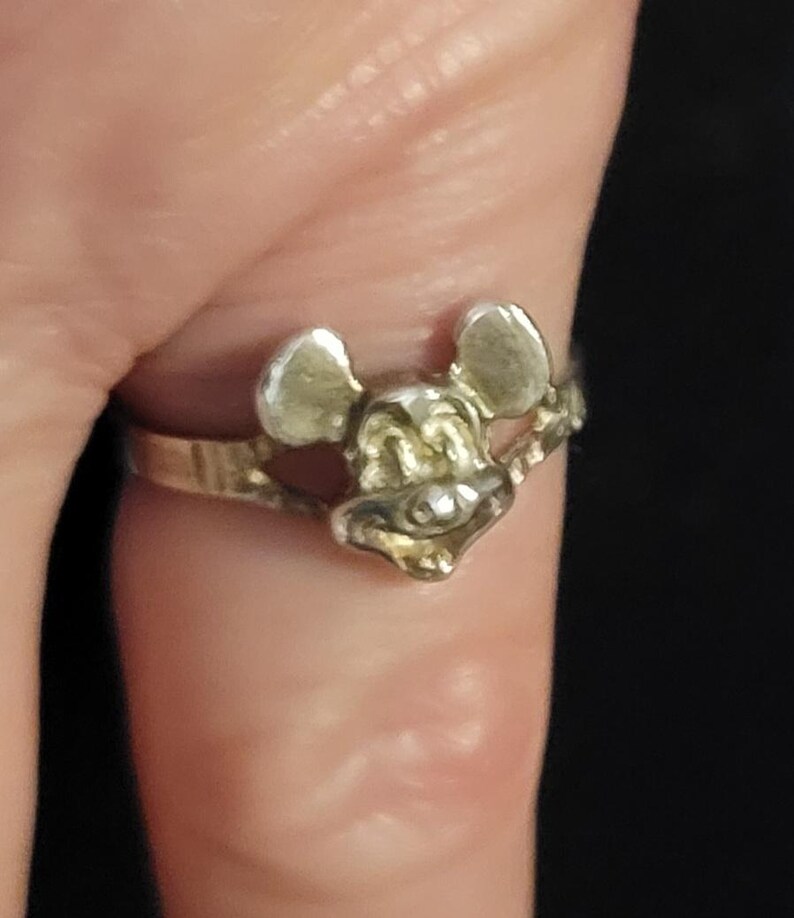 Puede incluir: Un anillo plateado con un dise&ntilde;o de la cara de Mickey Mouse. El anillo tiene una banda delgada, y la cara de Mickey Mouse est&aacute; detallada con orejas, nariz y una boca sonriente. El anillo se lleva en un dedo.