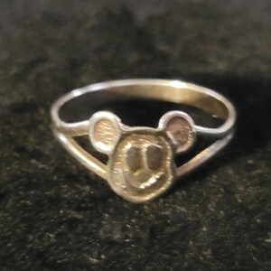 Puede incluir: Un anillo de metal plateado que presenta una cara circular central con dos peque&ntilde;as orejas redondeadas en la parte superior, similar a una cabeza de rat&oacute;n estilizada. La banda del anillo se divide en dos segmentos finos que se unen a los lados.