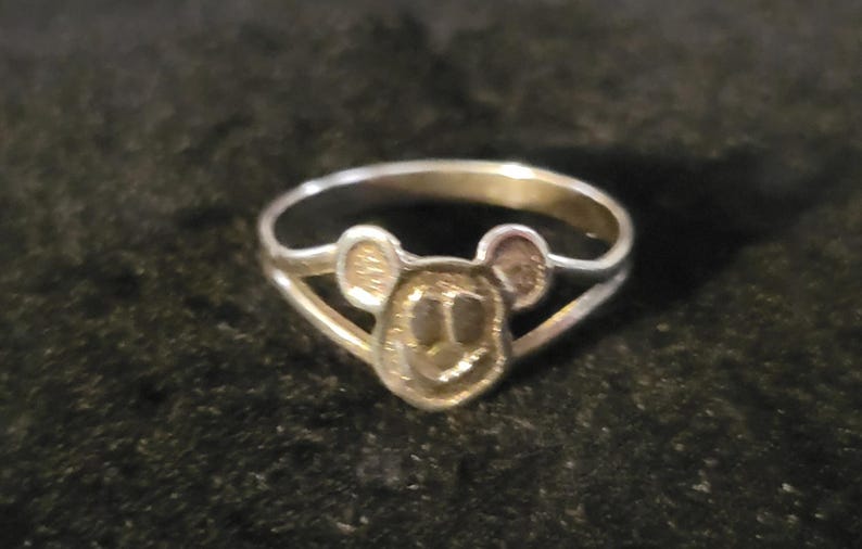 Puede incluir: Un anillo de metal plateado que presenta una cara circular central con dos peque&ntilde;as orejas redondeadas en la parte superior, similar a una cabeza de rat&oacute;n estilizada. La banda del anillo se divide en dos segmentos finos que se unen a los lados.