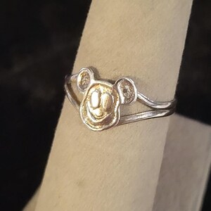 Puede incluir: Un anillo de metal plateado que presenta un emblema central con forma de cabeza de rat&oacute;n de dibujos animados con orejas redondeadas y una cara sonriente. La banda del anillo se divide en dos hilos de metal plateado finos y paralelos.