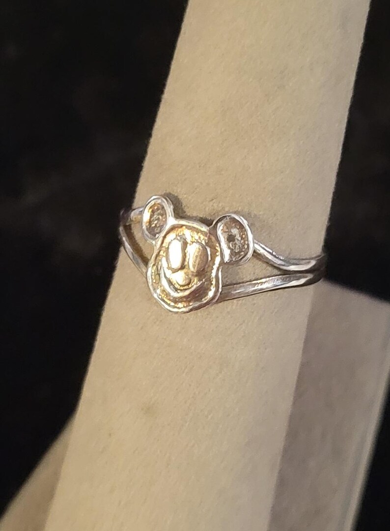 Puede incluir: Un anillo de metal plateado que presenta un emblema central con forma de cabeza de rat&oacute;n de dibujos animados con orejas redondeadas y una cara sonriente. La banda del anillo se divide en dos hilos de metal plateado finos y paralelos.