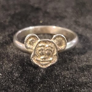 Pu&ograve; includere: Un anello color argento che presenta il volto modellato e in rilievo di un topo dei cartoni animati con grandi orecchie arrotondate. La fascia &egrave; liscia e circolare, collegata alla base del design della testa del topo. Metallo lucido.