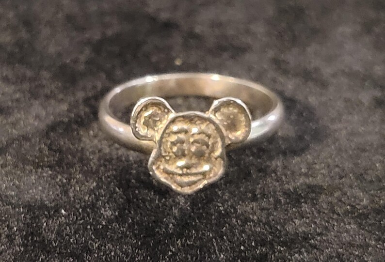 Pu&ograve; includere: Un anello color argento che presenta il volto modellato e in rilievo di un topo dei cartoni animati con grandi orecchie arrotondate. La fascia &egrave; liscia e circolare, collegata alla base del design della testa del topo. Metallo lucido.