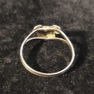 Puede incluir: Un anillo de metal en tono plateado con un dise&ntilde;o de v&aacute;stago dividido que converge en un elemento decorativo central. La banda es delgada y circular, mostrando un acabado met&aacute;lico pulido que refleja la luz en su estructura.